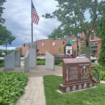 Photo of John Rich Park Veterans Memorial in summer. Green grass and background includes photo of YMCA on HWY 61