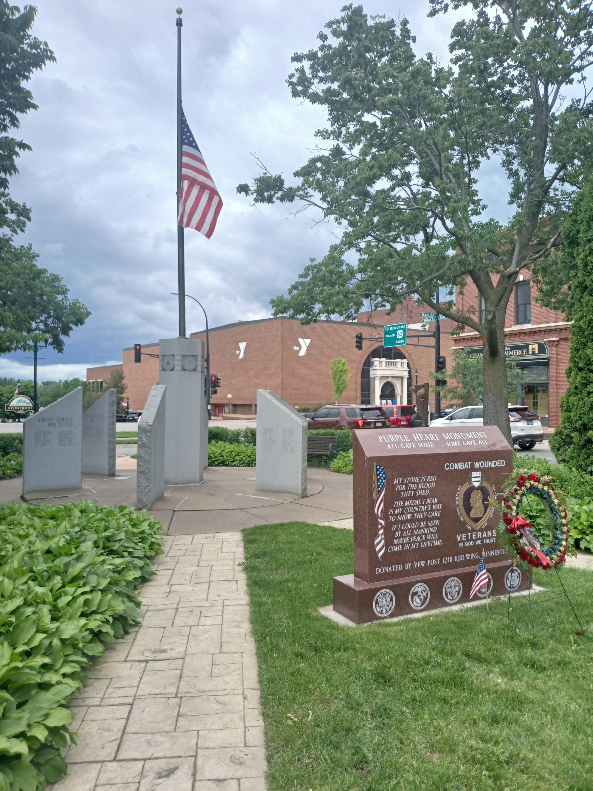 Photo of John Rich Park Veterans Memorial in summer. Green grass and background includes photo of YMCA on HWY 61